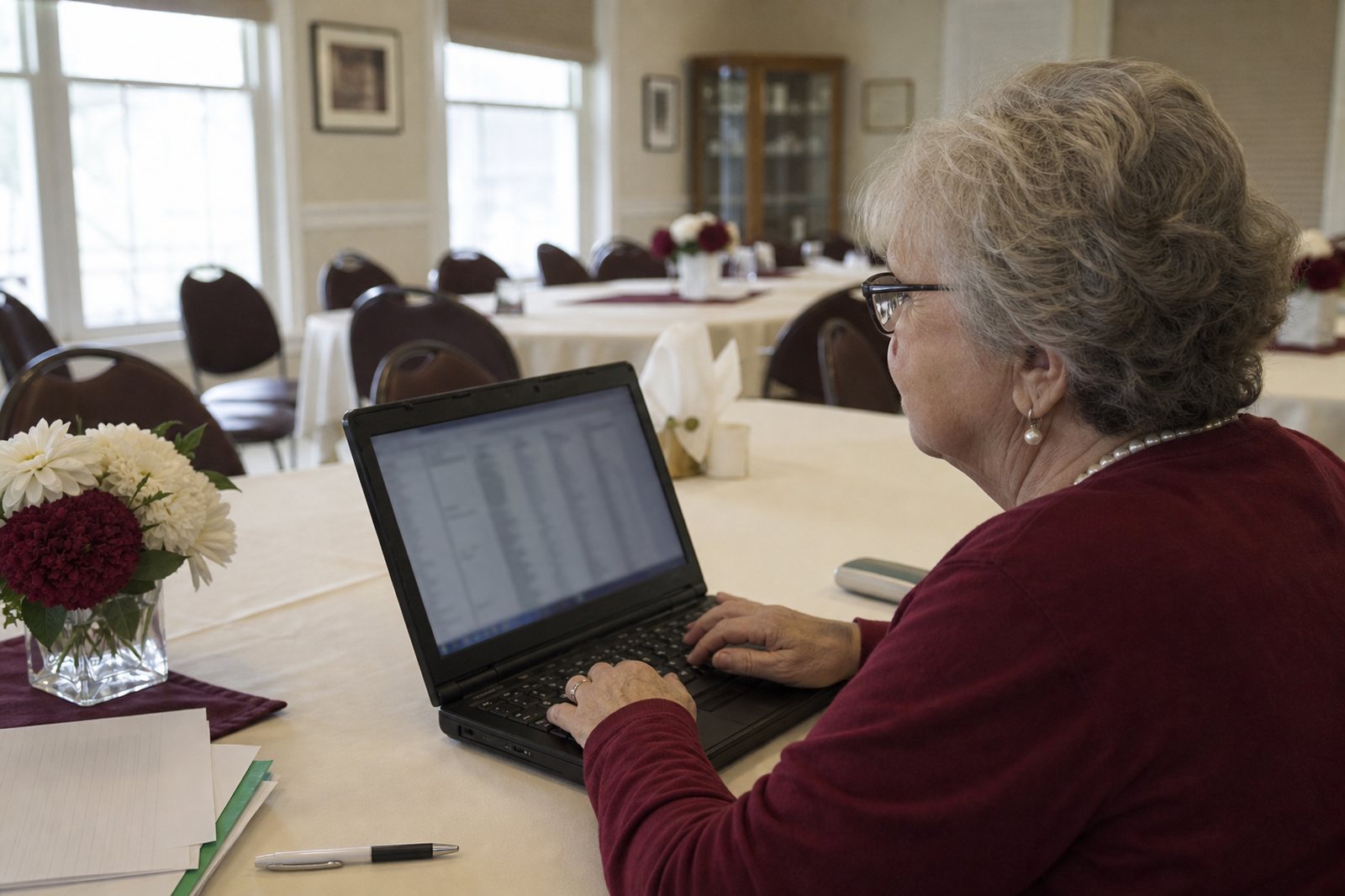 A chapter treasurer reviewing a membership roster on a laptop at a table in a bright meeting room with chairs set for a gathering